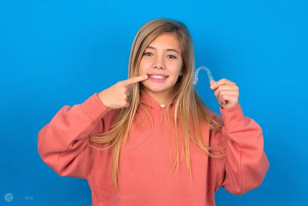 A girl in a pink sweater holding a clear aligner and pointing at her teeth, showing an effective method for how to fix overbite in Bakersfield and Tehachapi, CA.