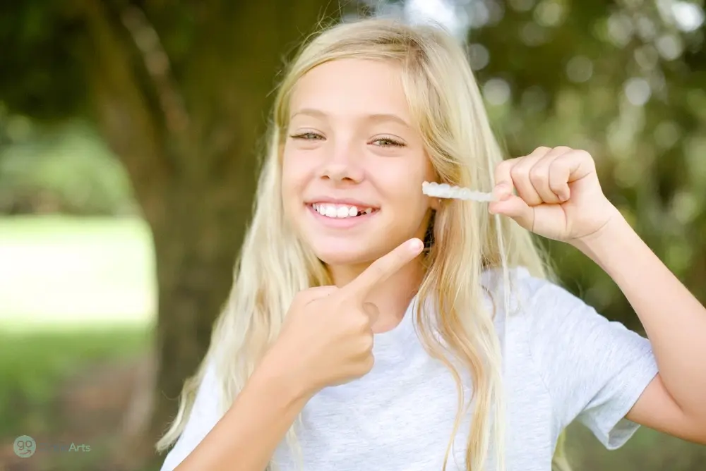 Blonde girl outdoors pointing at her braces with a big smile, highlighting orthodontic care for kids not knowing Does Insurance Cover Invisalign Bakersfield and Tehachapi, CA