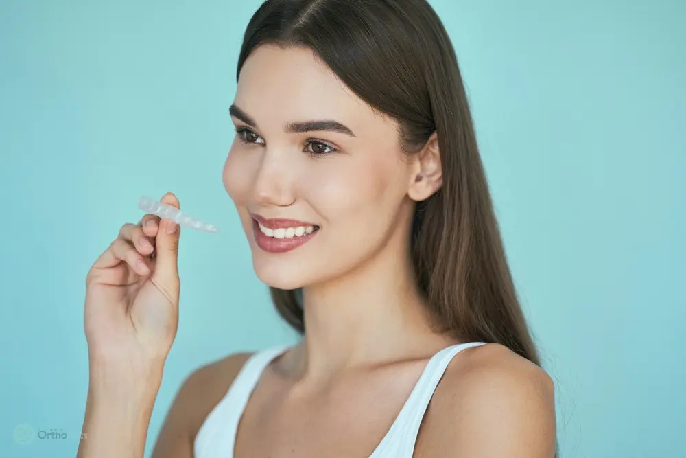Smiling woman with straight dark hair holding a clear aligner tray in front of a blue background, illustrating Does Insurance Cover Invisalign Bakersfield and Tehachapi, CA