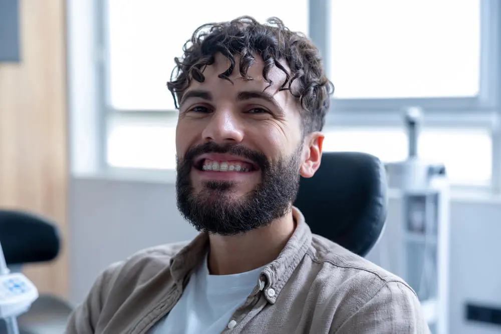 A man with curly hair and a beard smiles in a dental chair, showing clear braces at Gianquinto Orthodontic Arts in Tehachapi, CA