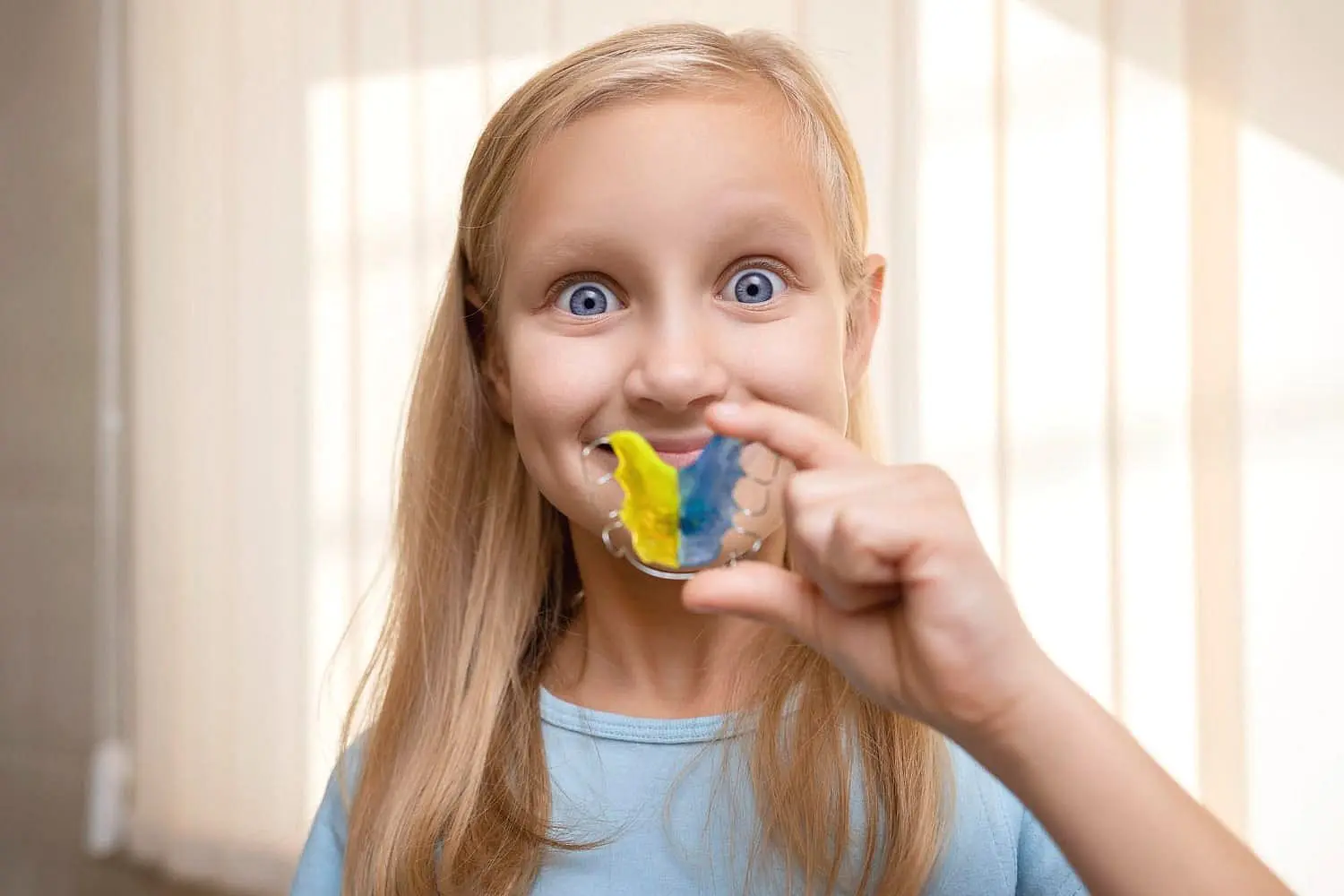 Smiling, a young girl holds a yellow and blue retainer after her visit with Children’s Orthodontist at Gianquinto Orthodontic Arts in Bakersfield, Tehachapi or Mammoth Lakes, CA.