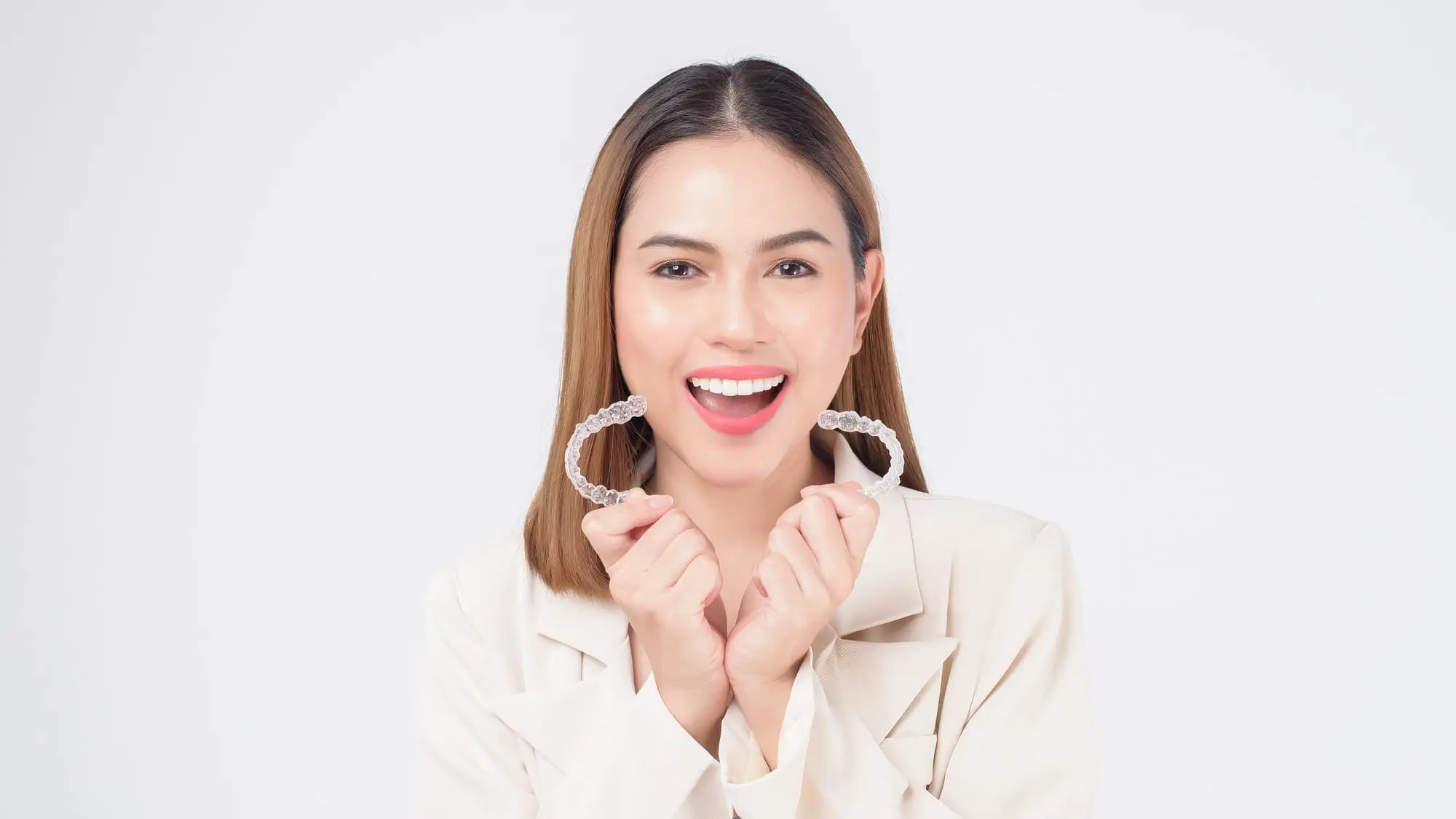 A woman in a white blazer smiles, holding two Invisalign clear aligners, representing Gianquinto Orthodontic Arts in Mammoth Lakes, CA.