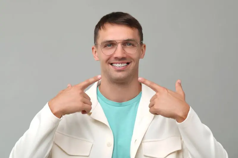A man in glasses and a white jacket points to his metal braces, smiling, for Gianquinto Orthodontic Arts in Tehachapi, CA