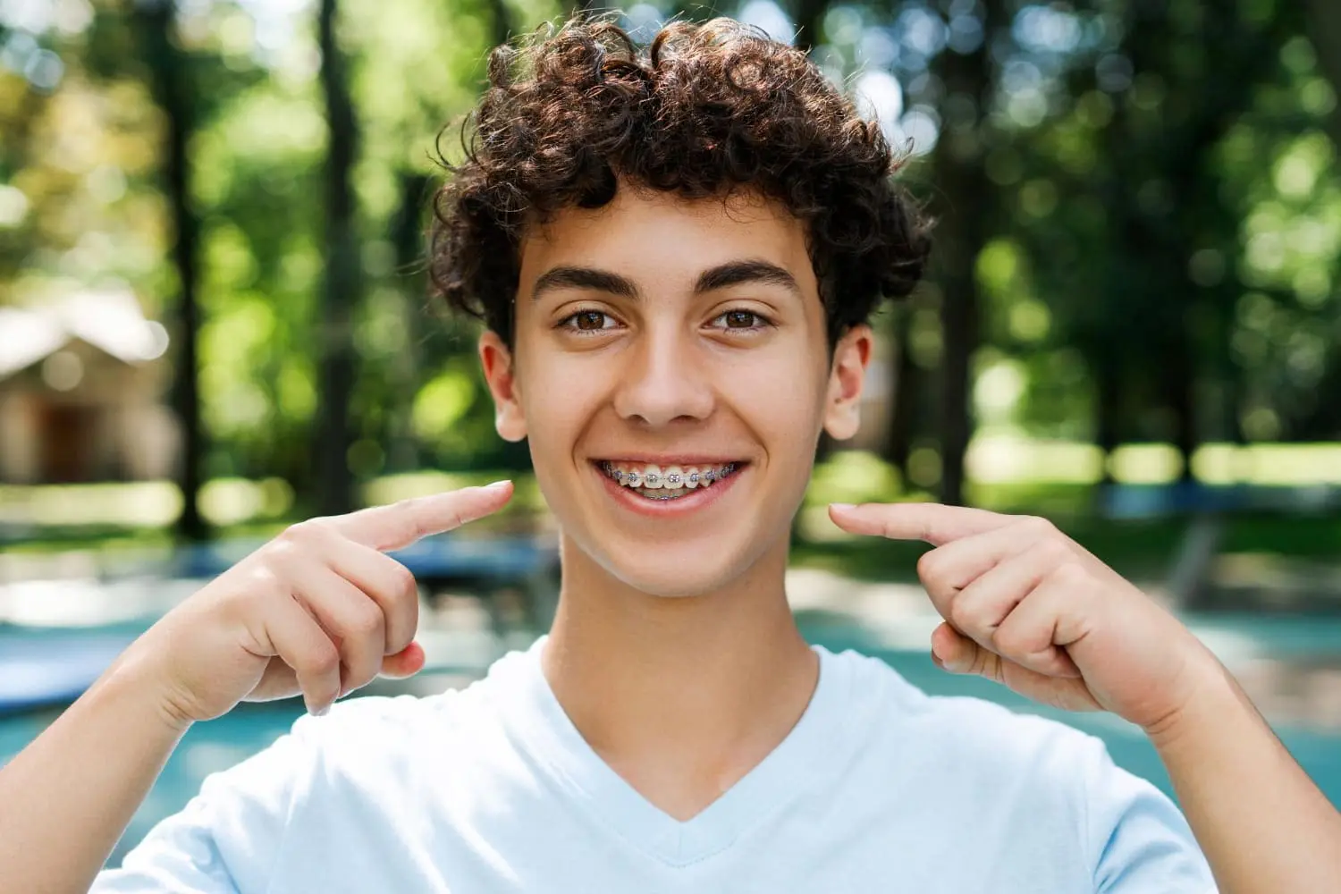 A teenage boy smiles and points to his metal braces outdoors for underbite treatment, promoting Gianquinto Orthodontic Arts in Bakersfield, Tehachapi and Mammoth Lakes, CA.