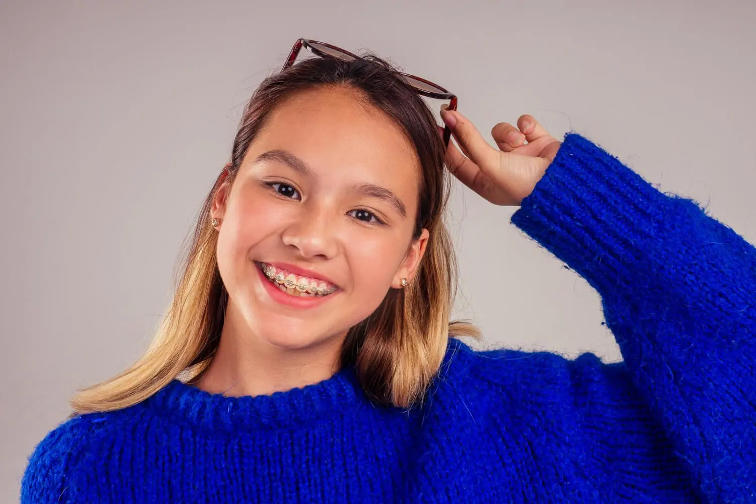 A smiling young person in a blue sweater and braces, holding eyeglasses above their head at Gianquinto Orthodontic Arts in Mammoth Lakes, CA.