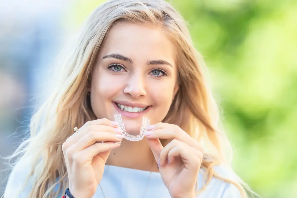 Smiling young woman with long blonde hair holds an Invisalign aligner at Gianquinto Orthodontic Arts in Bakersfield, CA