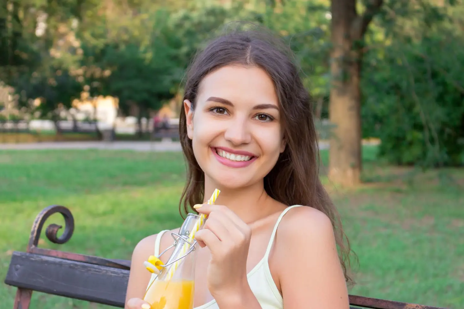 A young woman on a park bench smiles, holding orange juice, reflecting the best orthodontics for Adults at Gianquinto Orthodontic Arts in Bakersfield, Tehachapi or Mammoth Lakes, CA.