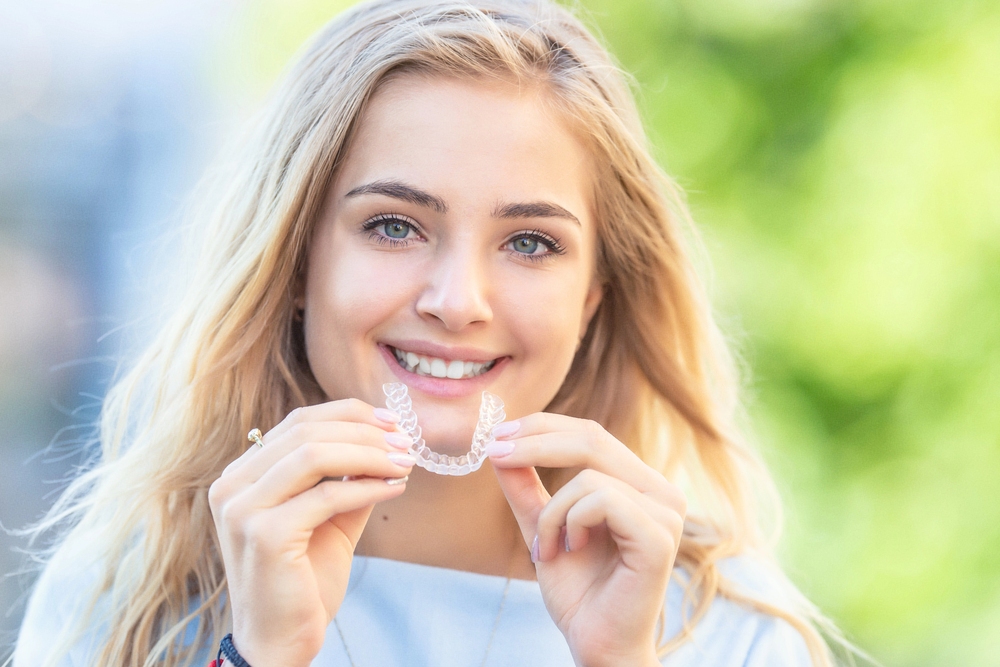 a blond girl smiling while holding invisalign clear aligner close to her mouth, wondering about Invisalign cost in Bakersfield and Tehachapi, CA