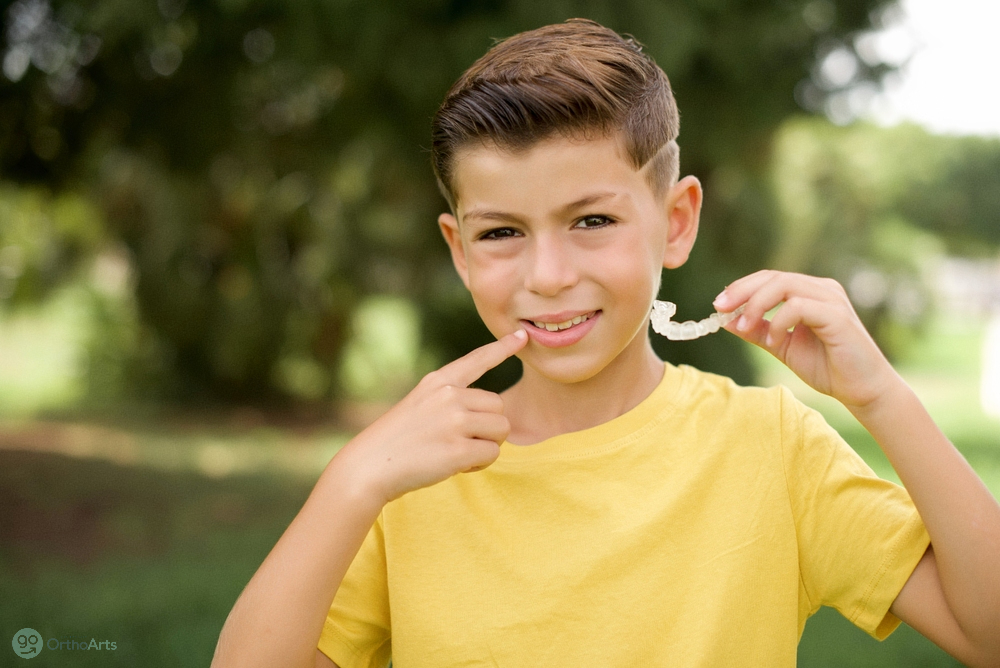 a boy smiling and pointing at his teeth while holding invisalign clear aligner close to his mouth, wondering about Invisalign cost in Bakersfield and Tehachapi, CA